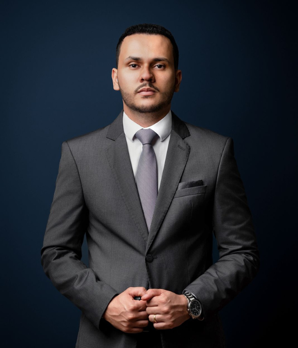 Retrato profissional de advogado criminalista em terno cinza escuro, postura confiante, braços cruzados, fundo neutro cinza, iluminação de estúdio, fotografia corporativa realista